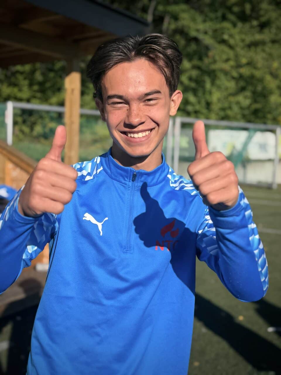 NTG-elev fotball bærum smiler til kamera med tomler opp. Han har på en blå genser med puma og NTG-logo. Bak han er det fotballmål og trær